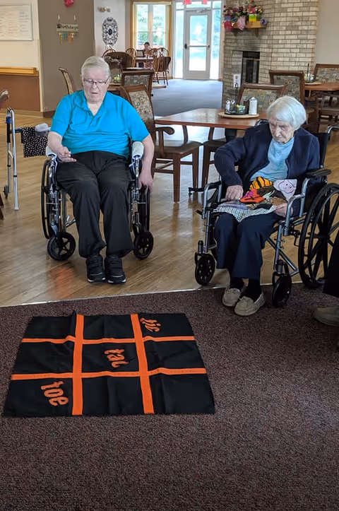 Two elderly individuals in wheelchairs playing a large floor tic-tac-toe game in a common area with tables, chairs, and a fireplace in the background.