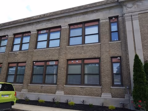 Exterior front of a two-story brick building facade with rows of windows, stone trim, a small planted bed, and a parked green car.