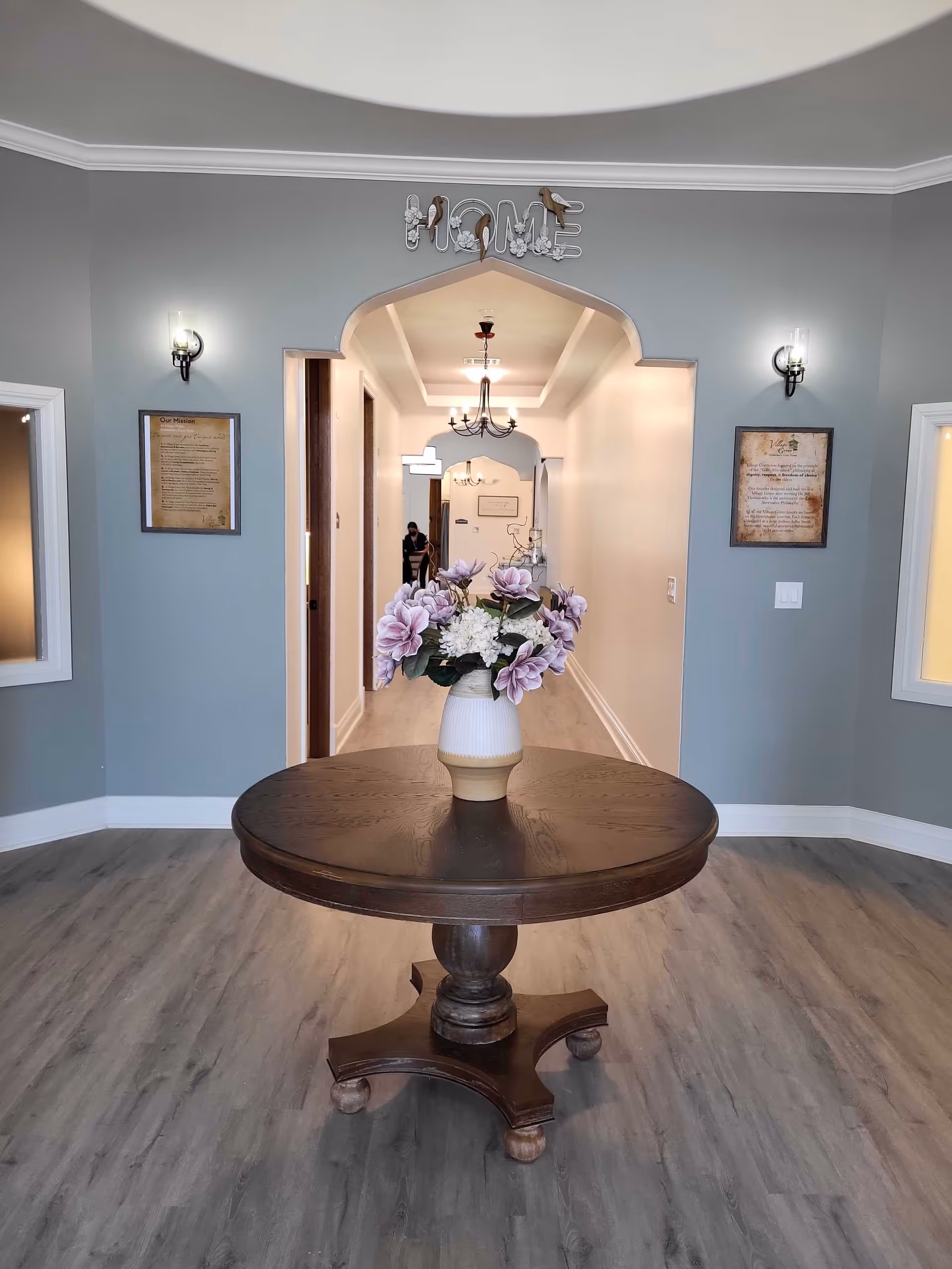 Round wooden table with a vase of pink flowers in a foyer leading to an arched hallway with chandeliers and a decorative "HOME" sign above the doorway.
