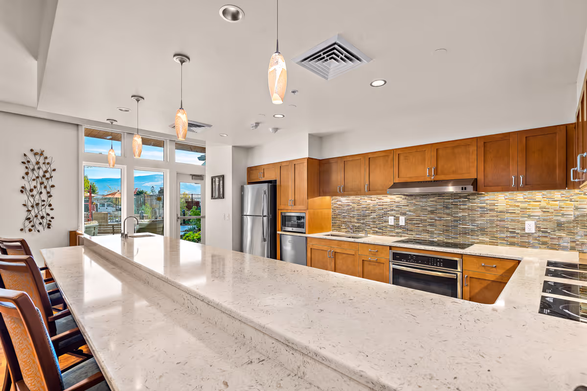 Bright modern kitchen featuring a long marble island with bar stools, pendant lights, wood cabinets, stainless steel appliances and glass doors to an outdoor area.