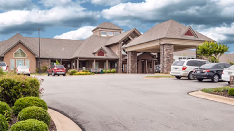Exterior view of Providence Assisted Living of Searcy building with a covered entrance, several parked cars, landscaped bushes, and a cloudy sky overhead.