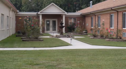 Outdoor courtyard area of Pine View Assisted Living Facility featuring a central fountain surrounded by landscaped flower beds with rose bushes, bordered by brick and beige buildings with windows and a central entrance.