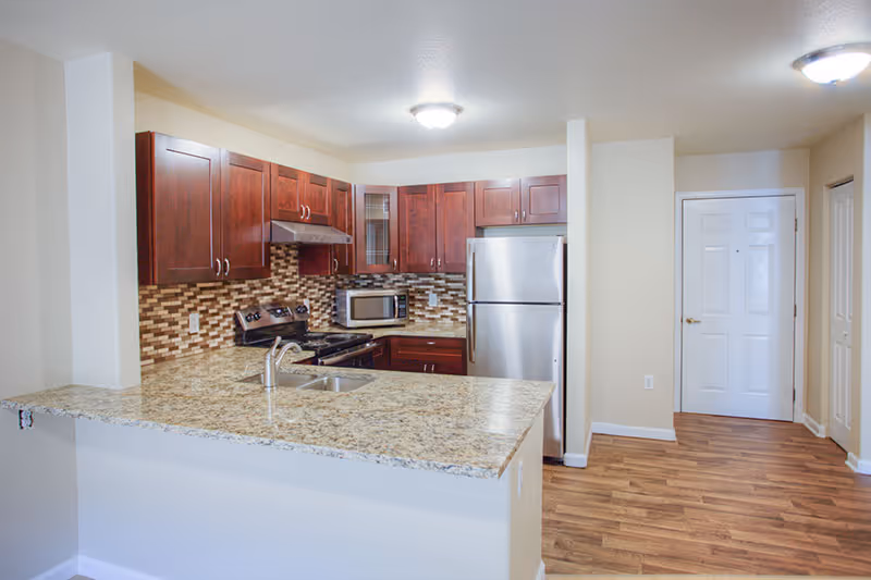 Interior view of a kitchen area in a senior living facility featuring granite countertops, a stainless steel refrigerator, stove, microwave, wooden cabinets, and a tiled backsplash. The kitchen opens to a hallway with a white door and wood flooring.