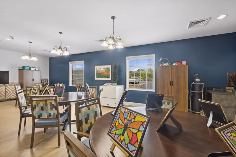 A bright and spacious room with multiple round wooden tables and chairs featuring colorful patterned cushions. Each table displays framed artwork, including a prominent butterfly painting. The walls are painted blue with two windows letting in natural light, and there are cabinets and decorative items along the walls.