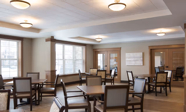 A bright communal dining room with multiple wooden tables and chairs, large windows, and ceiling lights.