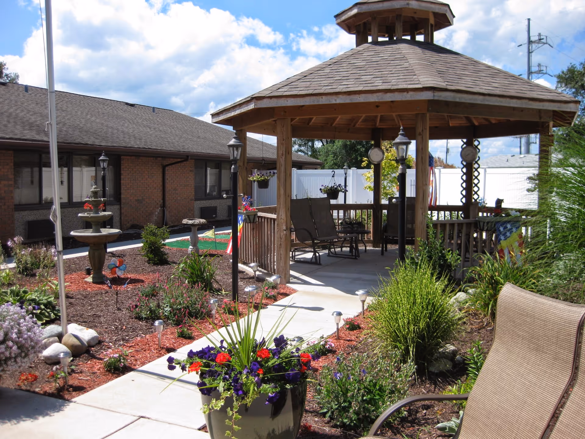 Outdoor garden area at East Lake Nursing and Rehabilitation Center featuring a wooden gazebo with seating, surrounded by landscaped flower beds, a small fountain, and a paved walkway under a partly cloudy sky.
