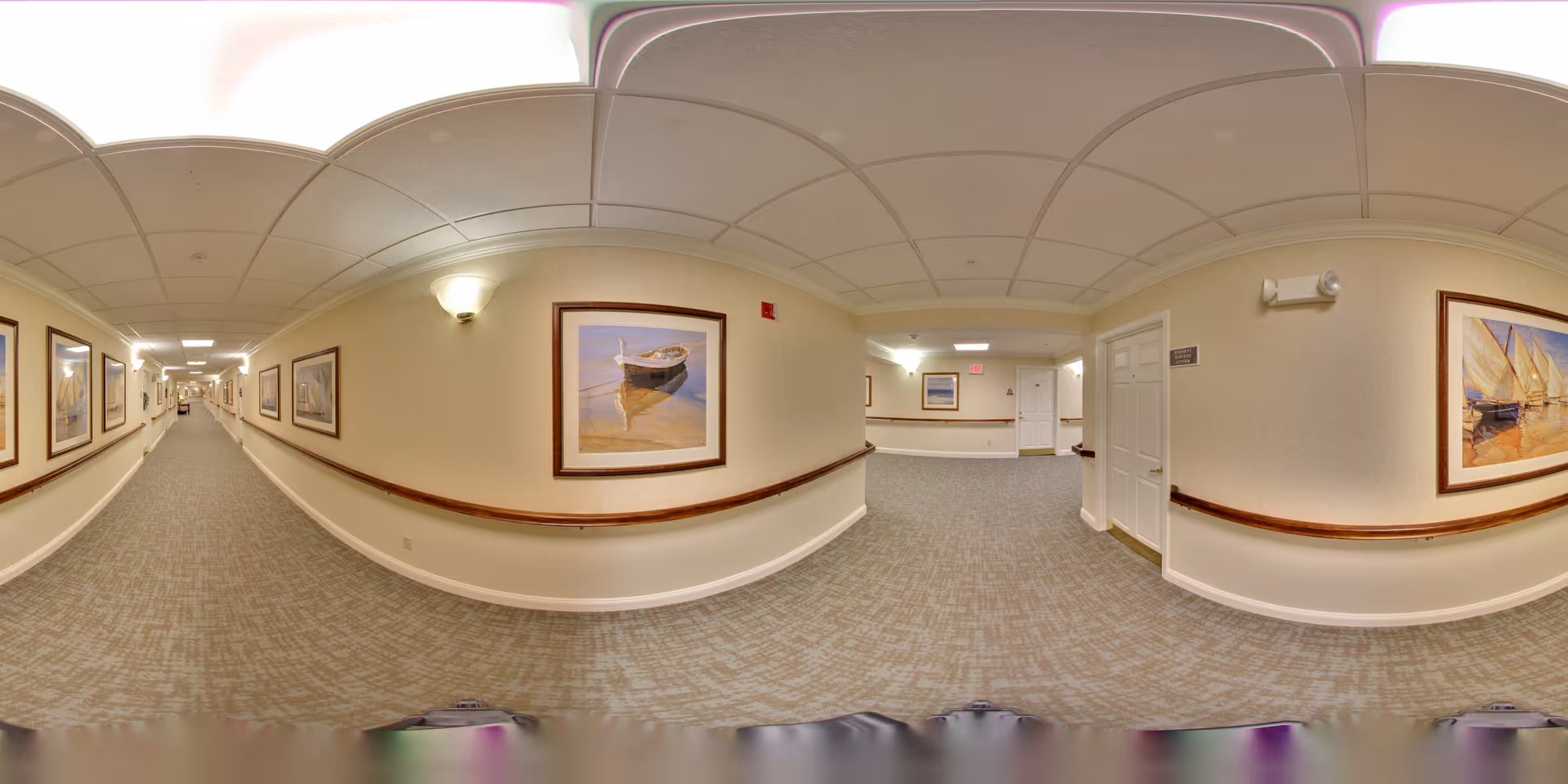 A long, carpeted hallway in a senior living facility with beige walls adorned with framed paintings of boats and nautical scenes. The ceiling has recessed lighting and a drop ceiling design. There are wooden handrails along the walls and several doors leading to rooms. The hallway is well-lit and clean.