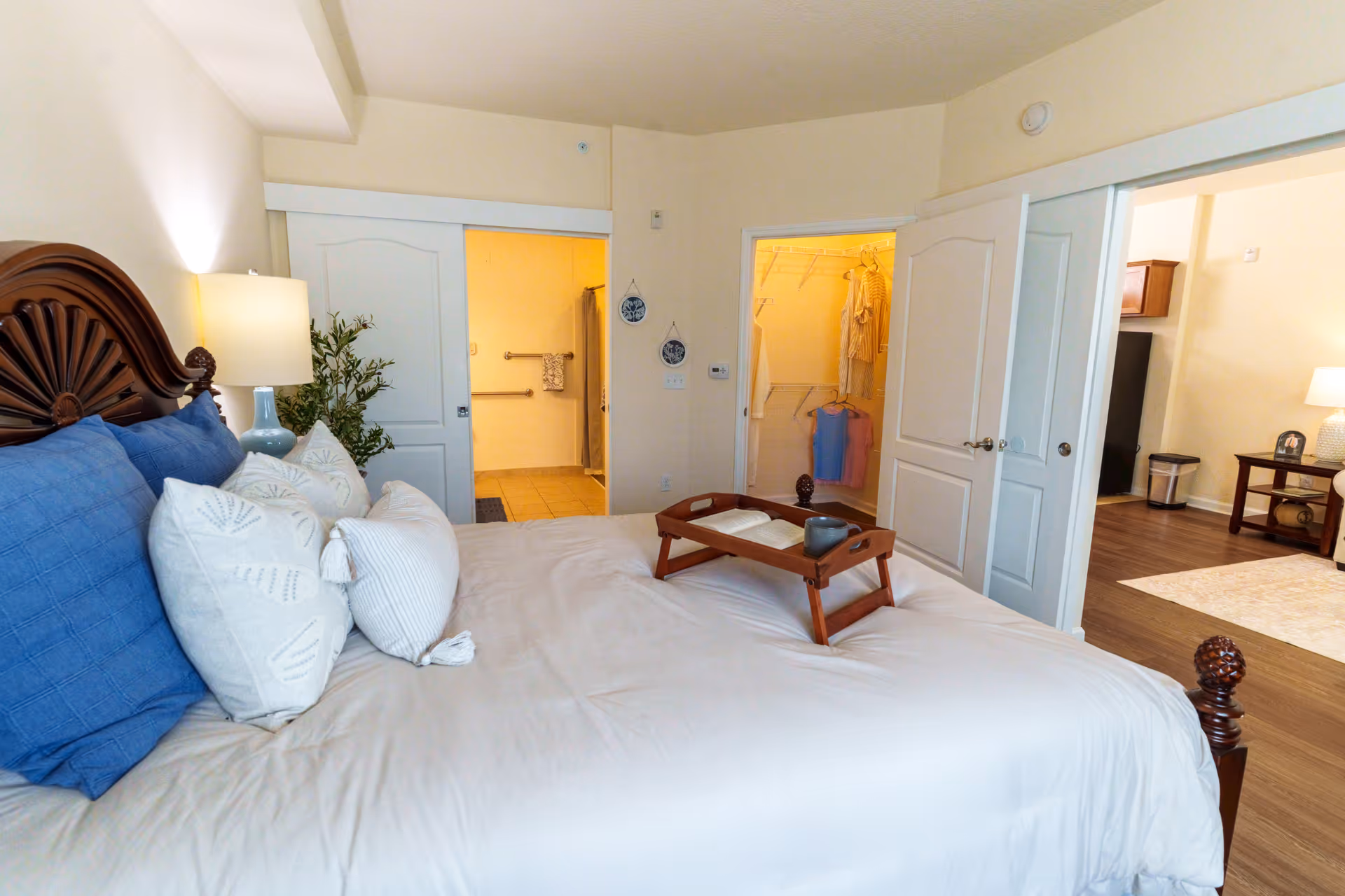Well-appointed bedroom with a made bed, decorative pillows and a breakfast tray, with open doors showing a closet and adjacent living area.