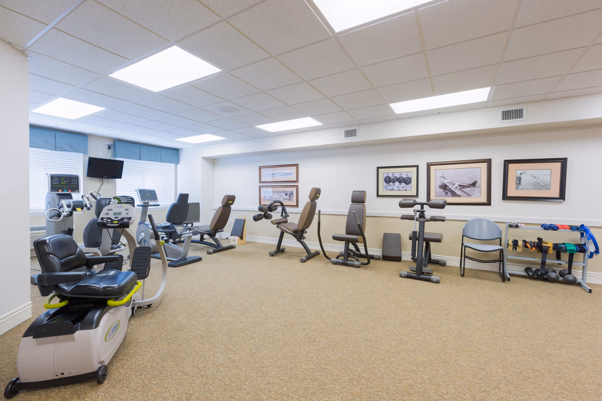 A well-lit exercise room in a senior living facility featuring various fitness equipment including recumbent bikes, weight machines, and free weights. The room has beige carpeting, white walls with framed pictures, and windows with blue valances allowing natural light.