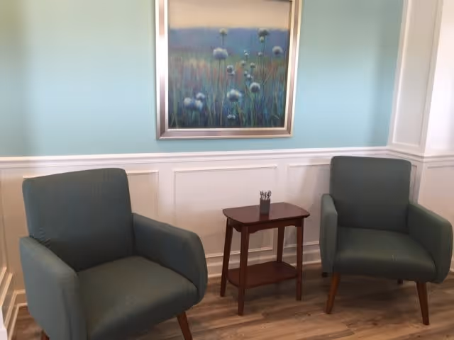 Two teal upholstered armchairs with wooden legs are positioned on either side of a small wooden side table with a container holding pens or pencils. The background features a light blue wall with white wainscoting and a framed painting of flowers in soft colors hanging above the chairs.