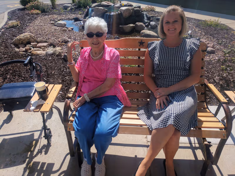 An elderly woman wearing sunglasses, a pink shirt, and blue pants sits on a wooden bench next to a younger woman in a black and white patterned dress. They are outdoors near a small rock water feature and garden area. A walker with a cup and tissue on its tray is positioned next to the bench.