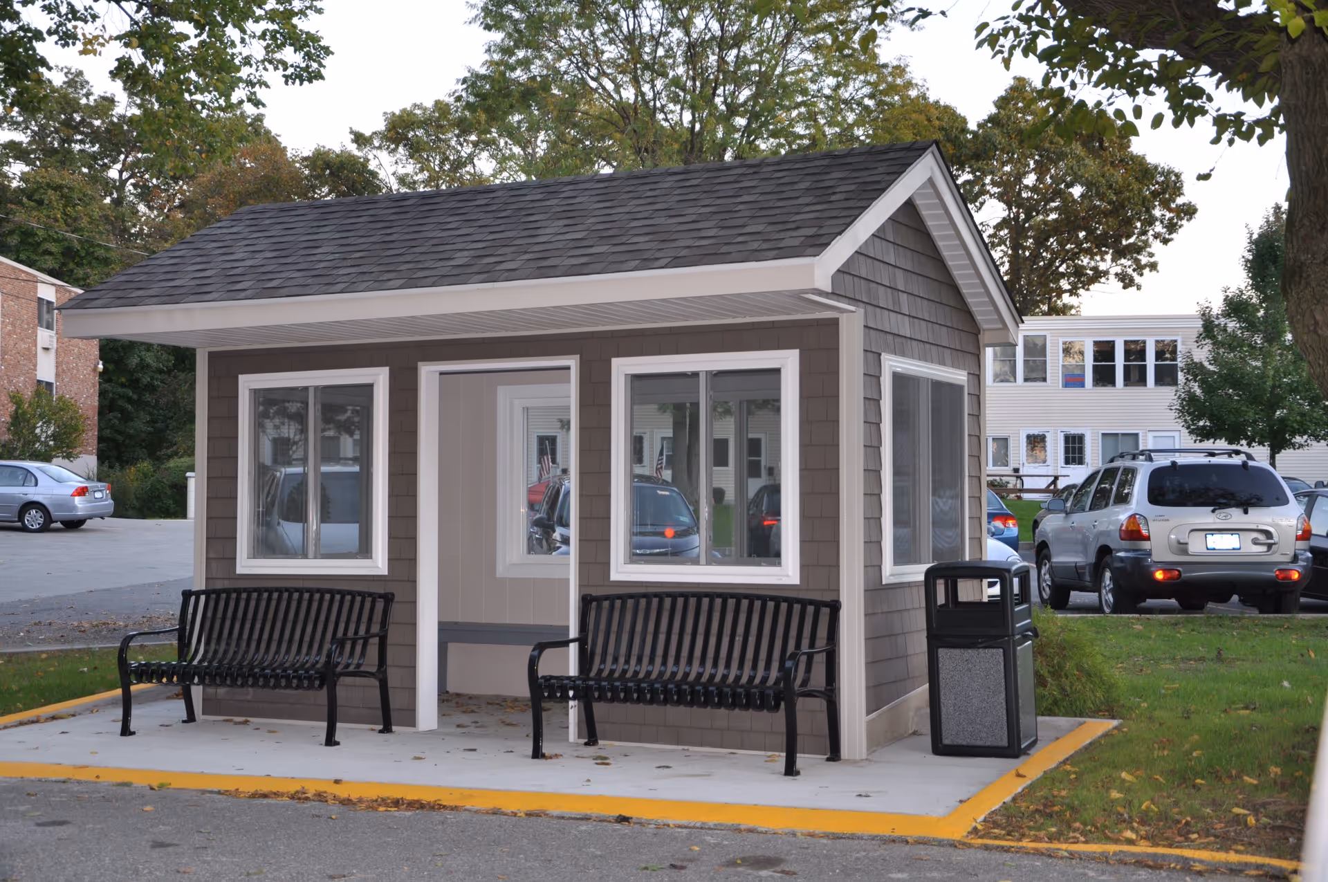 A small gray shelter with a shingled roof and white trim, featuring two black metal benches on either side of an open doorway. The shelter is situated on a concrete pad with a yellow painted edge, next to a trash can. In the background, there are parked cars and residential buildings surrounded by trees.
