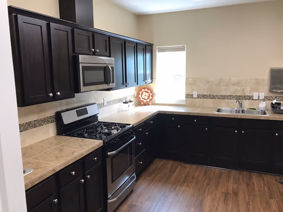 Kitchen with dark wood cabinets, stainless steel stove and microwave, tiled countertops and a double sink by a window.