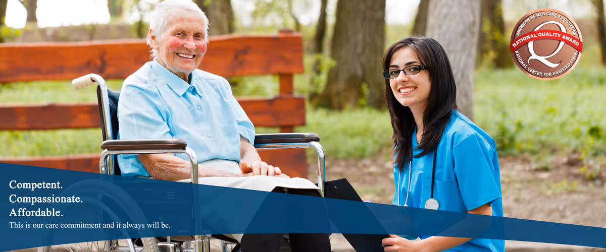 An elderly woman sitting in a wheelchair outdoors next to a young female healthcare worker in blue scrubs who is smiling and holding a clipboard. They are in a park-like setting with trees and a wooden bench in the background.