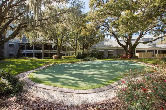 Outdoor putting green surrounded by trees and landscaping at Regency Oaks Health Center, with buildings visible in the background under a clear sky.
