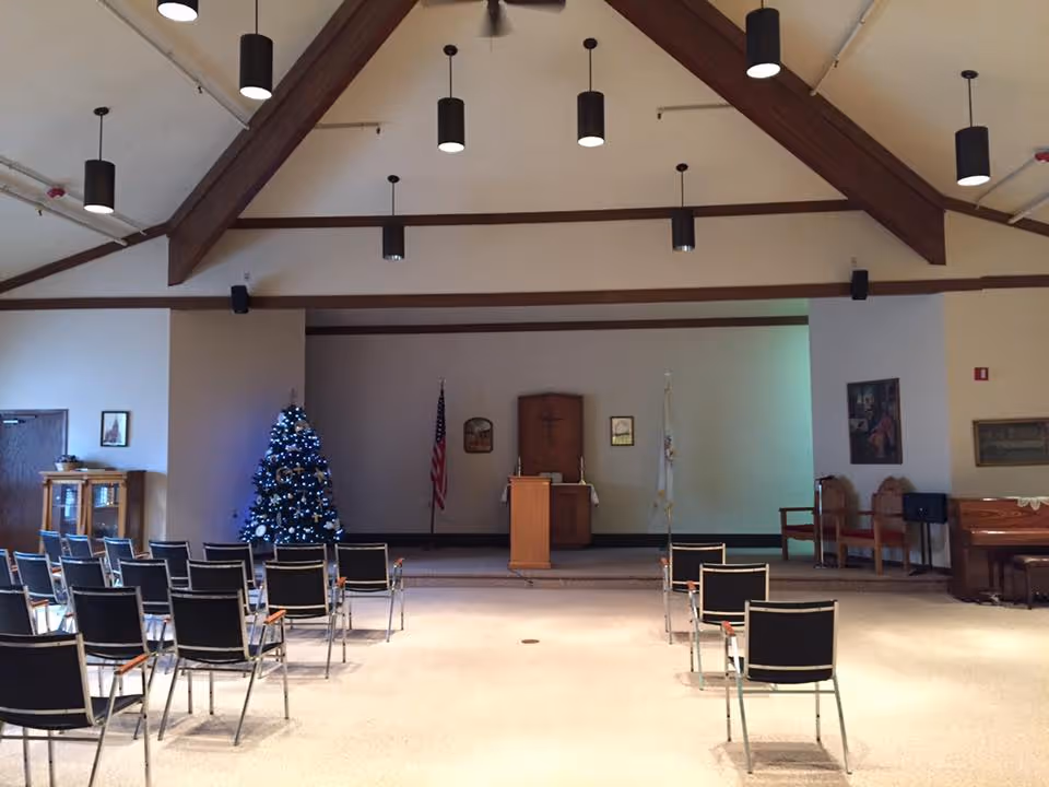 Interior view of a chapel or meeting room with rows of black chairs facing a wooden podium on a raised platform. Behind the podium are an American flag, a cross mounted on a wooden panel, and another flag. A decorated Christmas tree is visible to the left side of the platform. The ceiling has exposed wooden beams and multiple hanging cylindrical light fixtures.