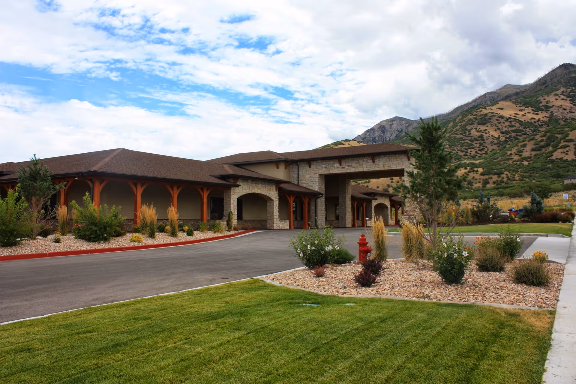 Exterior view of Seasons of Santaquin facility showing a single-story building with a covered entrance, surrounded by landscaped greenery, shrubs, and a lawn, with mountains in the background under a partly cloudy sky.