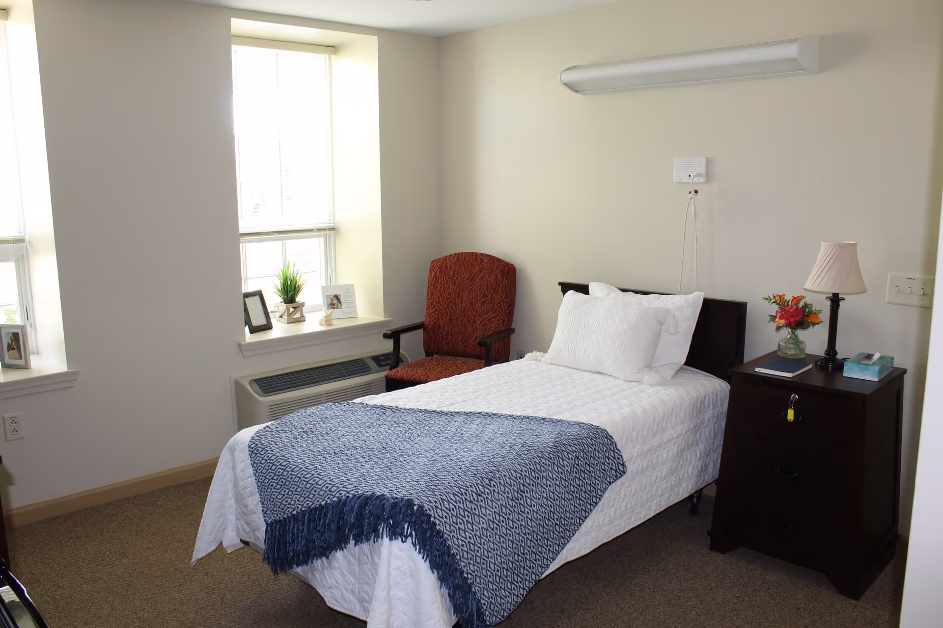 A neatly made single bed with white bedding and a blue patterned throw blanket in a senior living bedroom. Next to the bed is a dark wooden nightstand with a lamp, a small vase of flowers, a book, and a tissue box. A red upholstered chair is positioned near two windows with white blinds, and decorative items including framed photos and a small plant are placed on the window sill. The room has beige carpet and light-colored walls.