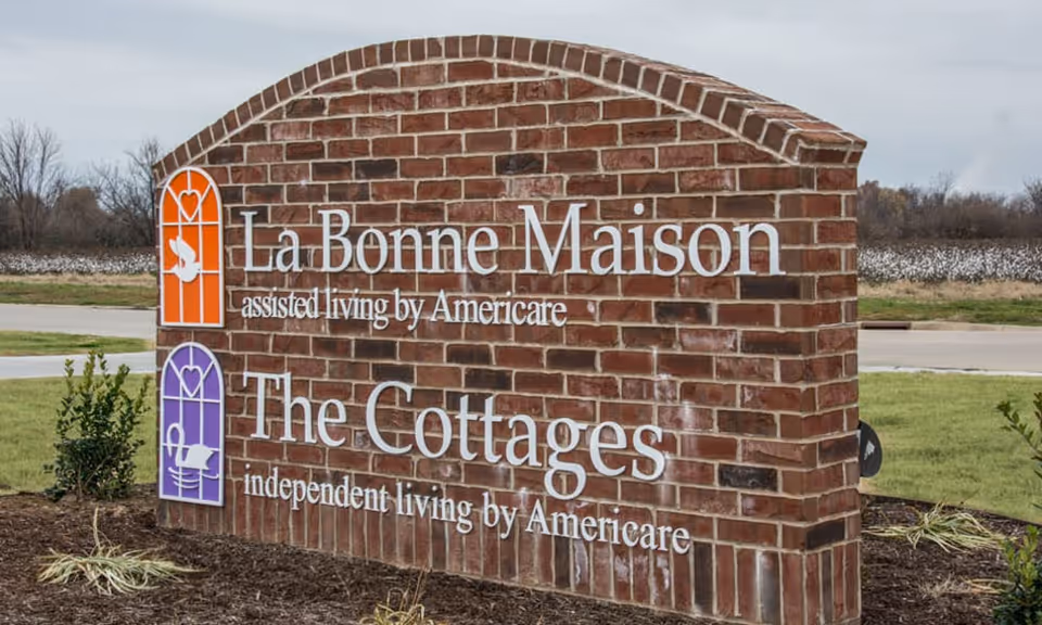 A large brick sign outdoors with white text displaying 'La Bonne Maison assisted living by Americare' and 'The Cottages independent living by Americare' with two colored icons, one orange and one purple, on the left side of the text. The sign is surrounded by grass and small plants with a road and trees in the background.