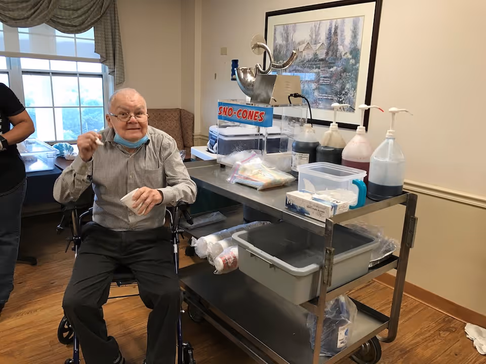 An elderly man sitting in a wheelchair inside a room, holding a cup and spoon, with a sno-cones machine and various syrup bottles on a metal cart beside him. The room has a window with curtains, a framed painting on the wall, and wooden flooring.