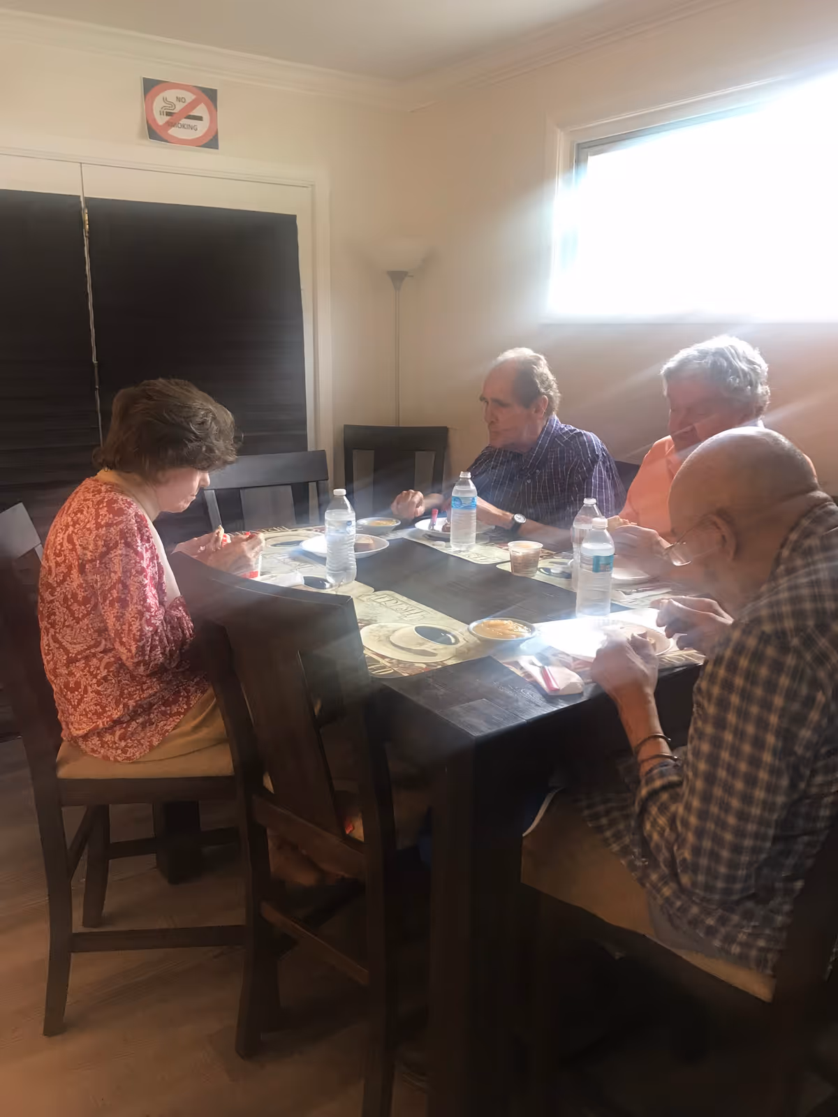 Four elderly people sitting around a dark wooden dining table eating a meal. The table has placemats, bottled water, and small bowls of food. The room has light-colored walls, a window with bright sunlight coming through, a floor lamp in the corner, and a 'No Smoking' sign above a door with closed black blinds.