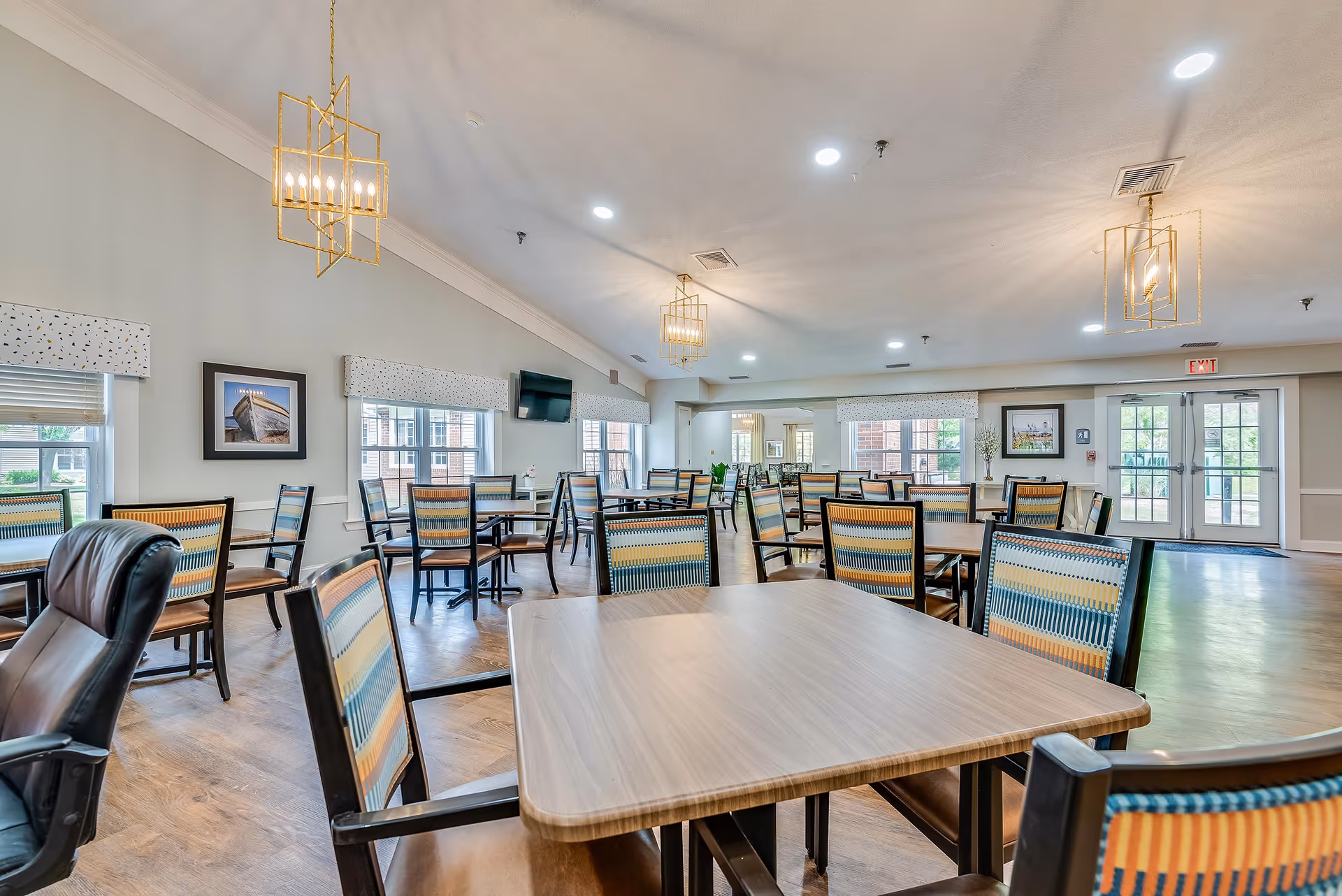 Spacious communal dining room with multiple tables and patterned chairs, large windows, and modern pendant lighting.