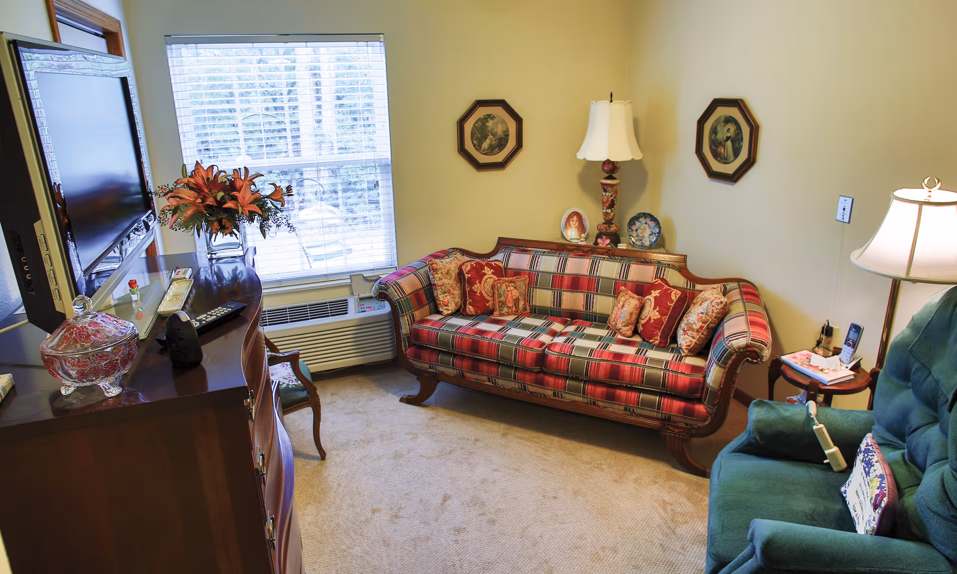 A cozy living room with a plaid upholstered sofa adorned with decorative pillows, a green armchair with a floral cushion, a wooden side table with a lamp and phone, a wooden dresser with a flat-screen TV mounted above it, a vase of flowers, and a window with blinds letting in natural light.
