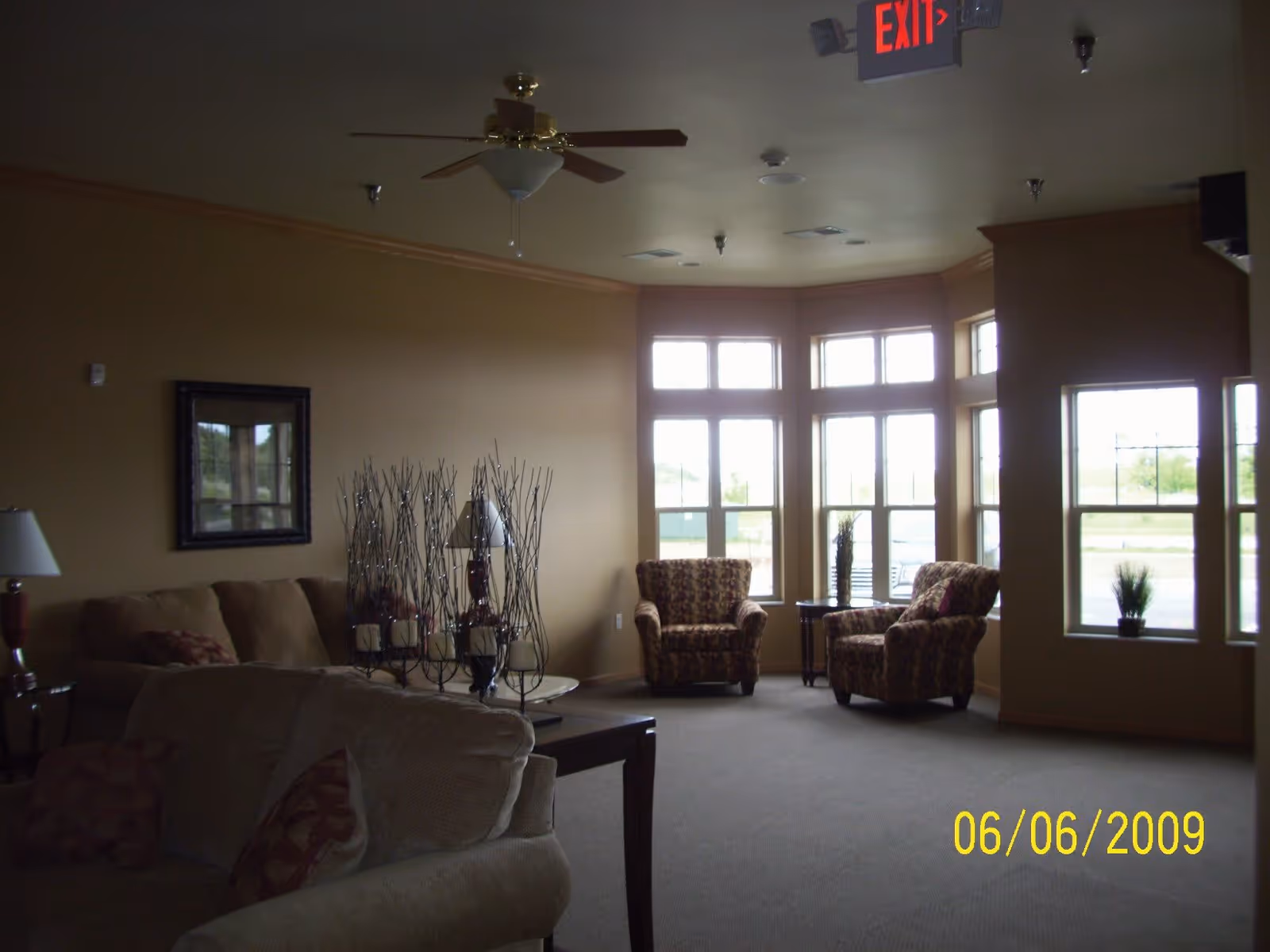 Spacious assisted-living lounge with sofas and patterned armchairs by large bay windows under a ceiling fan.