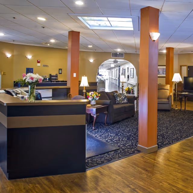 Interior view of a senior living facility lobby area with a reception desk on the left, decorated with a vase of flowers. The seating area includes a sofa, armchairs, side tables with lamps, and floral arrangements. The space has wooden flooring, patterned carpet, and warm lighting with ceiling lights and wall sconces. A hallway extends into the background through an arched doorway.