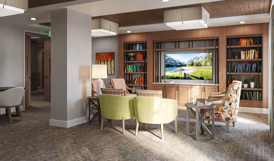 A cozy living room area in an assisted living facility featuring a large flat-screen TV mounted on a wooden wall unit with shelves filled with books and plants. The room has comfortable seating including two light green chairs, two patterned armchairs, and a small round wooden table. A lamp and a colorful painting are visible near a hallway entrance.