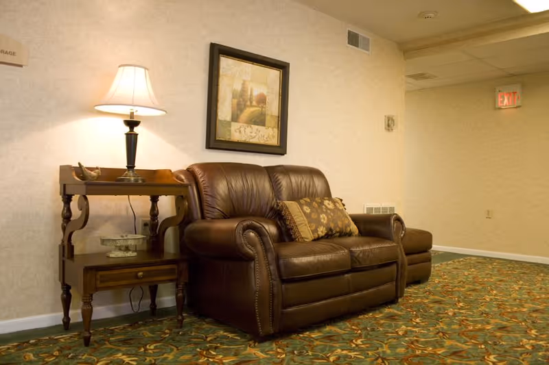 A cozy seating area with a brown leather loveseat adorned with two decorative pillows. Next to the loveseat is a wooden side table with a lamp and decorative items. A framed landscape painting hangs on the wall above the loveseat. The floor is covered with a patterned carpet, and an exit sign is visible in the background.