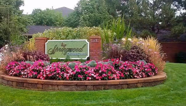 A landscaped garden area with a brick and stone raised flower bed filled with pink flowers and various green plants. In the center is a green sign with white trim that reads 'Springwood A Retirement Community'. Trees and shrubs are visible in the background.