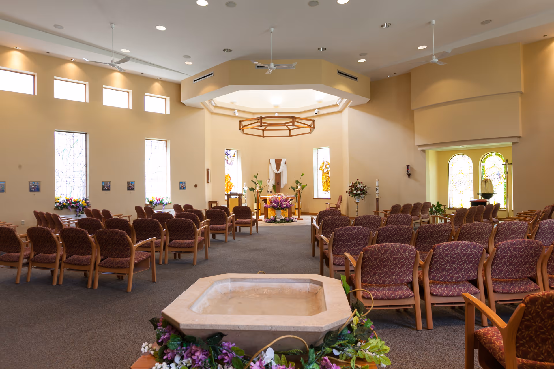 Sunlit chapel interior with rows of upholstered chairs facing an altar and stained glass windows.
