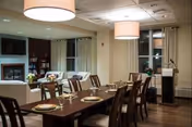 A dining room with a long wooden table set with plates, glasses, and cutlery. The room features large windows with curtains, two large ceiling lights, a fireplace, and a cozy seating area with white sofas and a bookshelf in the background.