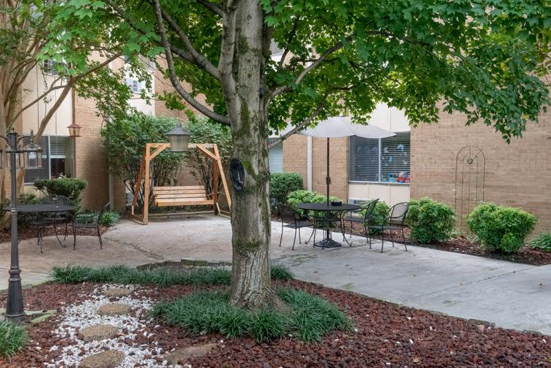 Outdoor courtyard area with a large tree in the center surrounded by landscaping with mulch and white stones. There is a wooden swing bench under a wooden frame, metal patio tables and chairs with umbrellas, and a brick building with windows in the background.