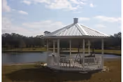 A white gazebo with a metal roof situated on a grassy area near a pond, with trees and a partly cloudy sky in the background.