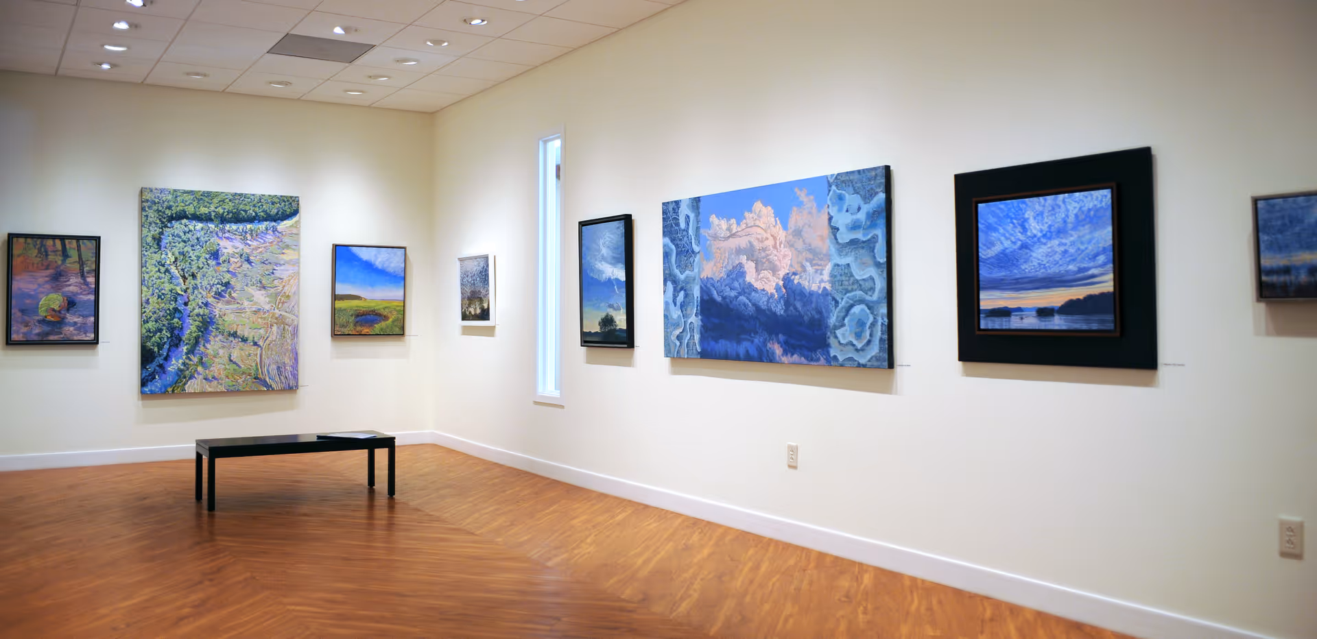 Interior view of an art gallery room with several landscape paintings hanging on white walls. The room has wooden flooring, a bench in the center, and recessed ceiling lights illuminating the artwork.