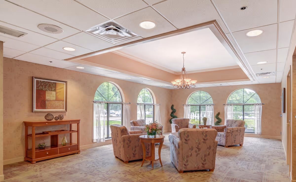 A bright and spacious living room area with four upholstered armchairs arranged around a small round wooden table with a flower arrangement. The room features large arched windows with sheer white curtains, allowing natural light to fill the space. The walls are decorated with light wallpaper and a framed abstract painting. A wooden console table with decorative items is placed against one wall. The ceiling has recessed lighting and a central chandelier.