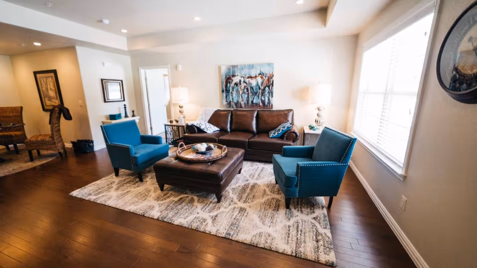 A cozy living room with a brown leather sofa, two blue armchairs, and a large ottoman on a patterned area rug. The room has hardwood floors, two table lamps on side tables, a large window with blinds, and a painting of horses on the wall.