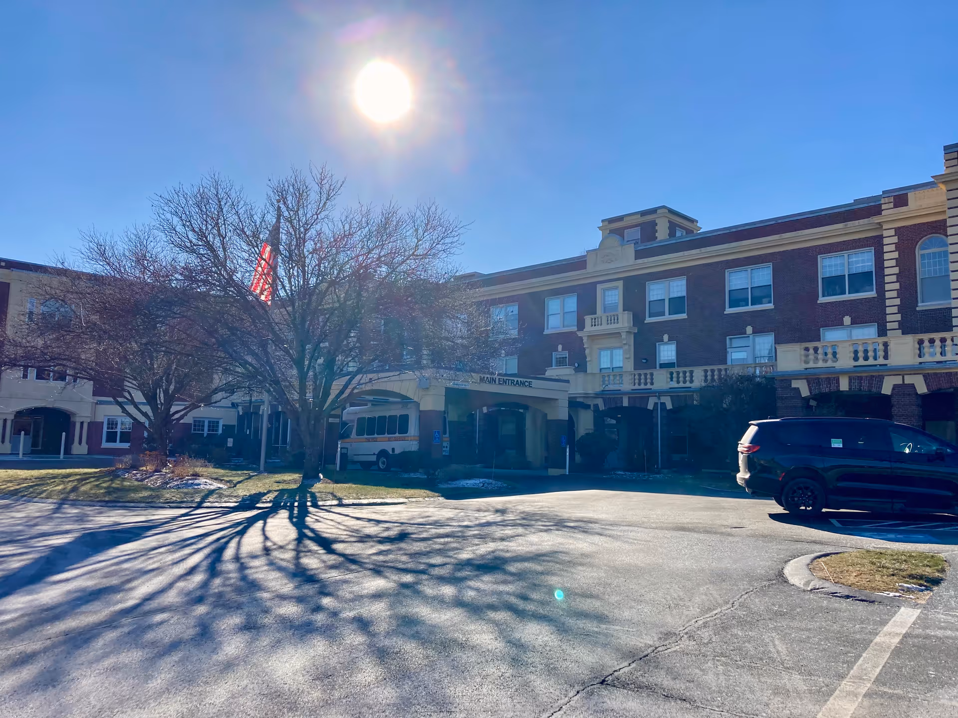 Exterior view of a multi-story brick building with a covered main entrance. There is a large leafless tree casting shadows on the driveway, an American flag near the entrance, a parked black vehicle, and a shuttle bus under the entrance canopy. The sun is shining brightly in a clear blue sky.
