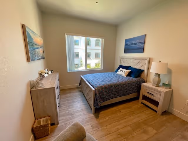 A bright bedroom with a wooden bed frame and blue bedding, a matching wooden dresser and nightstand, a lamp on the nightstand, a window with vertical blinds, and wall art above the bed and dresser.