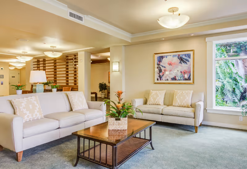 A bright and spacious living room area with two beige sofas adorned with patterned cushions, a wooden coffee table with a potted plant centerpiece, a large window showing green foliage outside, a framed floral painting on the wall, and soft ceiling lights illuminating the space.