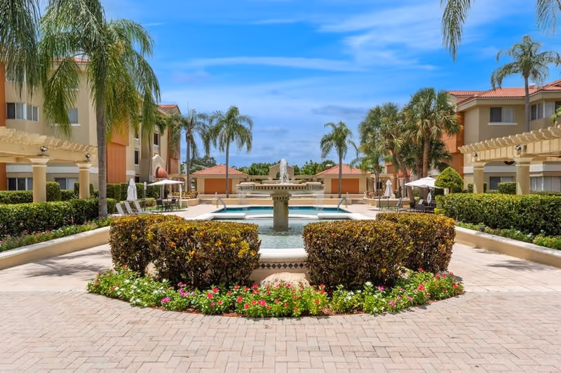 Outdoor courtyard area with a central water fountain surrounded by trimmed bushes and colorful flowers. Palm trees and two-story buildings with balconies and pergolas are visible under a bright blue sky with some clouds.