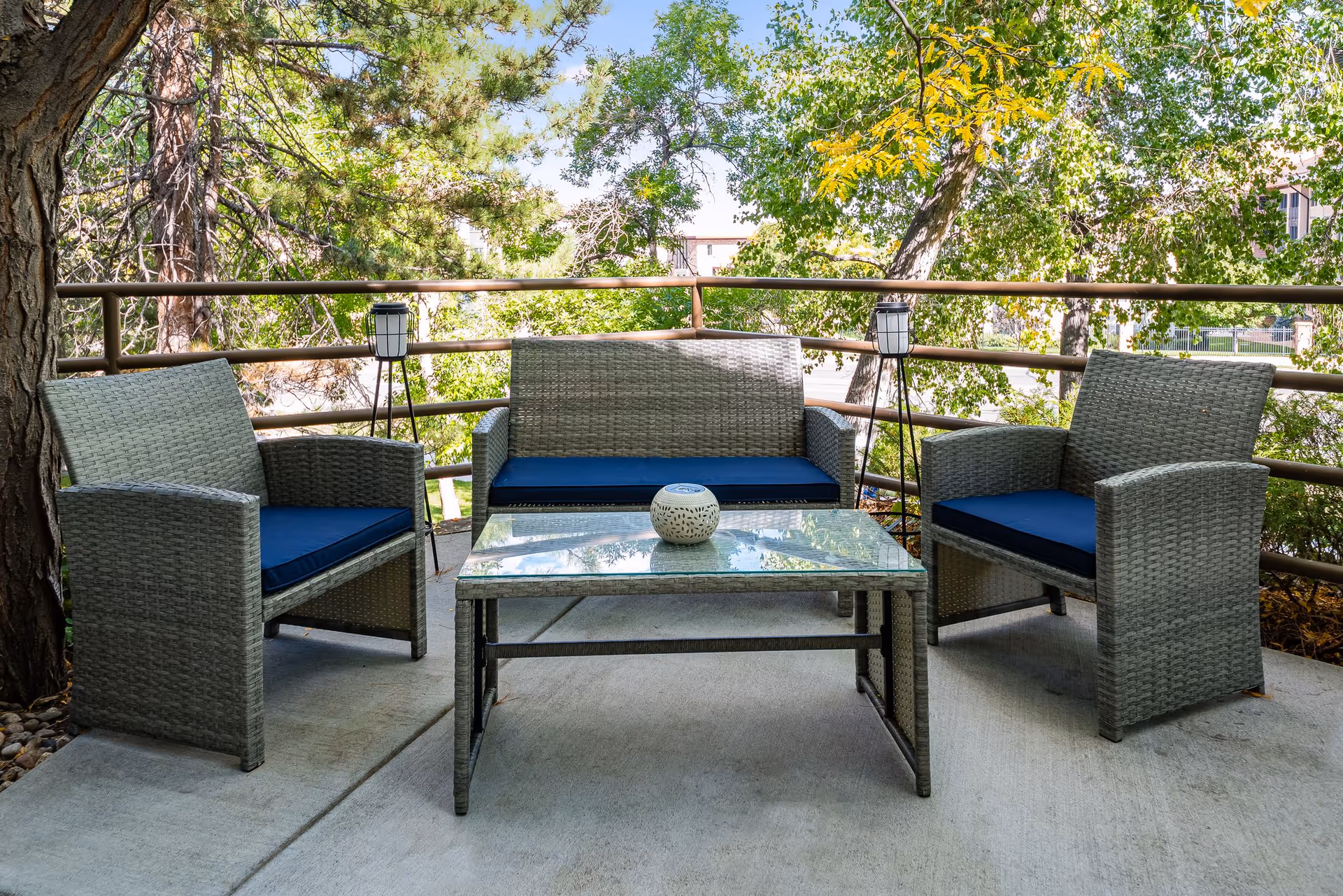 Outdoor patio seating with two wicker chairs and a loveseat with blue cushions arranged around a glass coffee table on a concrete patio surrounded by trees.