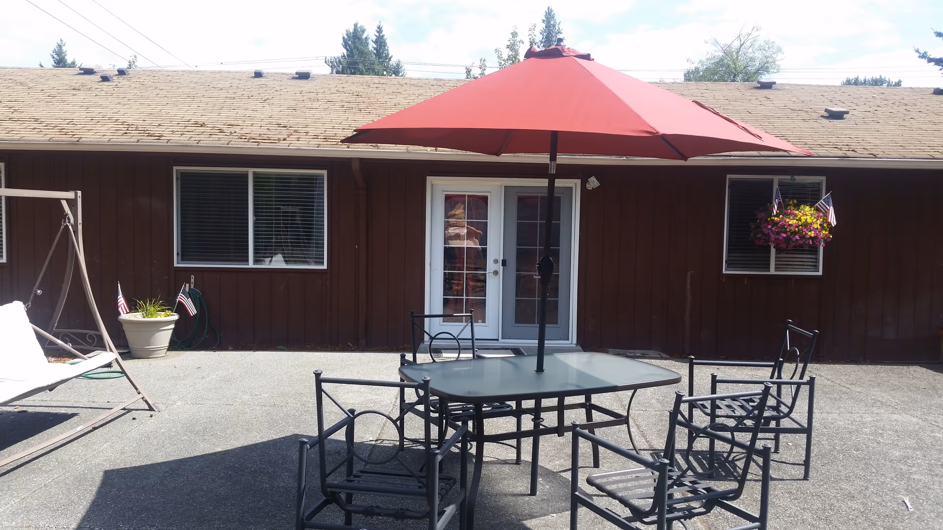 Outdoor patio area with a glass-top table and four metal chairs under a large red umbrella. There is a brown building with two windows and a glass door in the background. A hanging flower pot with colorful flowers and small American flags is visible on the right side, and a white swing with cushions is on the left side.