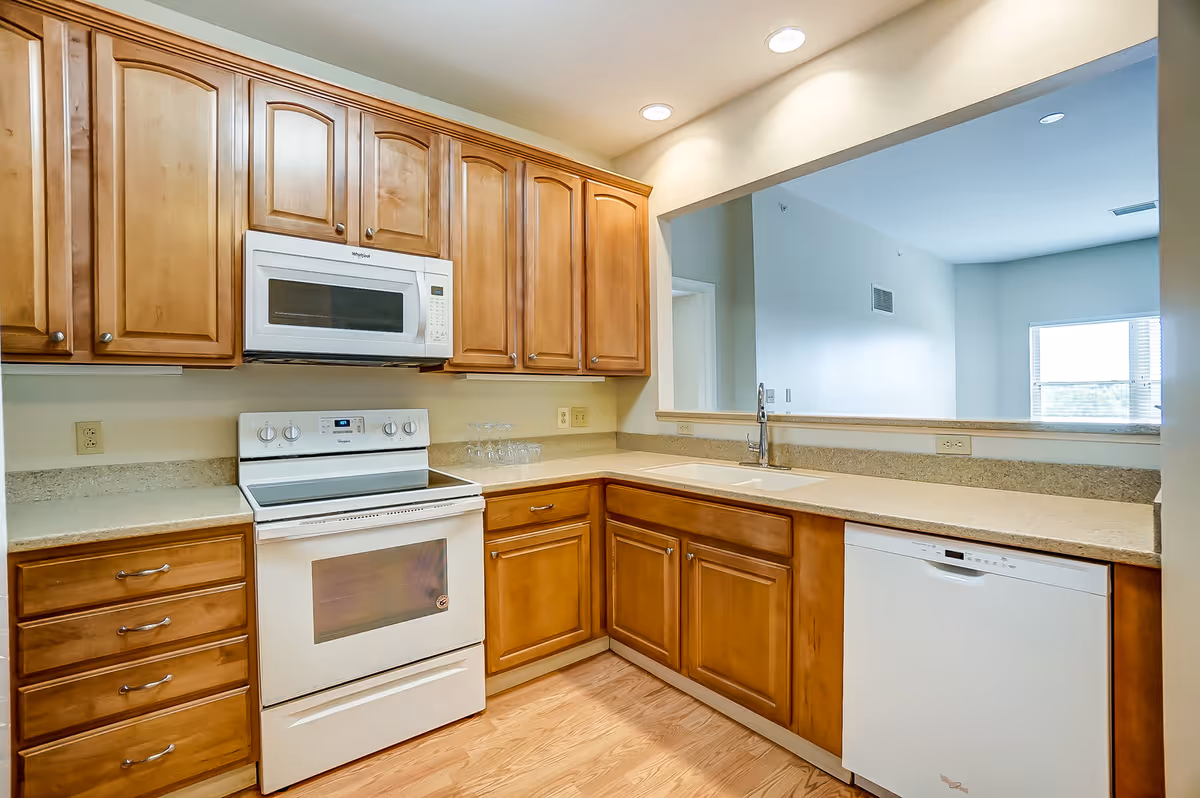 A kitchen with wooden cabinets, a white electric stove with oven, a white microwave above the stove, a white dishwasher, and a sink with a faucet. The kitchen has a light-colored countertop and wood flooring. There is a large pass-through window opening to an adjacent room with a window letting in natural light.