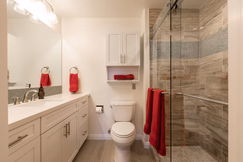 Clean modern bathroom with a white vanity, toilet, glass-enclosed tiled shower, and red towels.