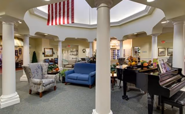 A spacious and well-lit common area in an assisted living facility featuring white columns, a floral armchair, a blue loveseat, a black grand piano with sheet music, and decorative plants. An American flag hangs from the ceiling, and the room has a carpeted floor with soft yellow walls and multiple doorways leading to other rooms.