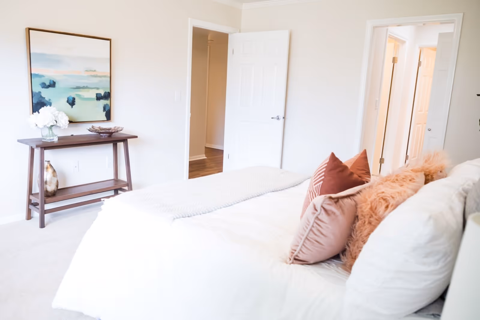 Bright bedroom with a white bed covered in multiple pillows in shades of pink and peach. A wooden console table against the wall holds a vase with white flowers and decorative items. A large abstract painting with blue and green tones hangs above the table. Two open doors reveal a hallway and another room with wooden flooring.
