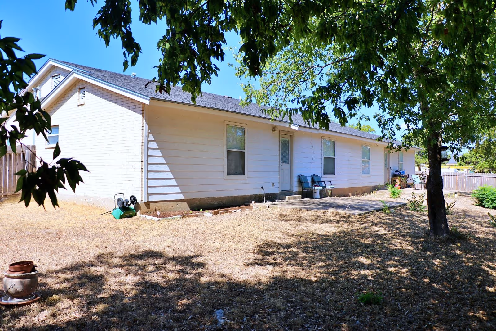 Exterior view of a single-story white house with a sloped roof, surrounded by a dry yard with sparse grass and a few trees providing shade. There are two chairs and some items near the back door on a small concrete patio area.